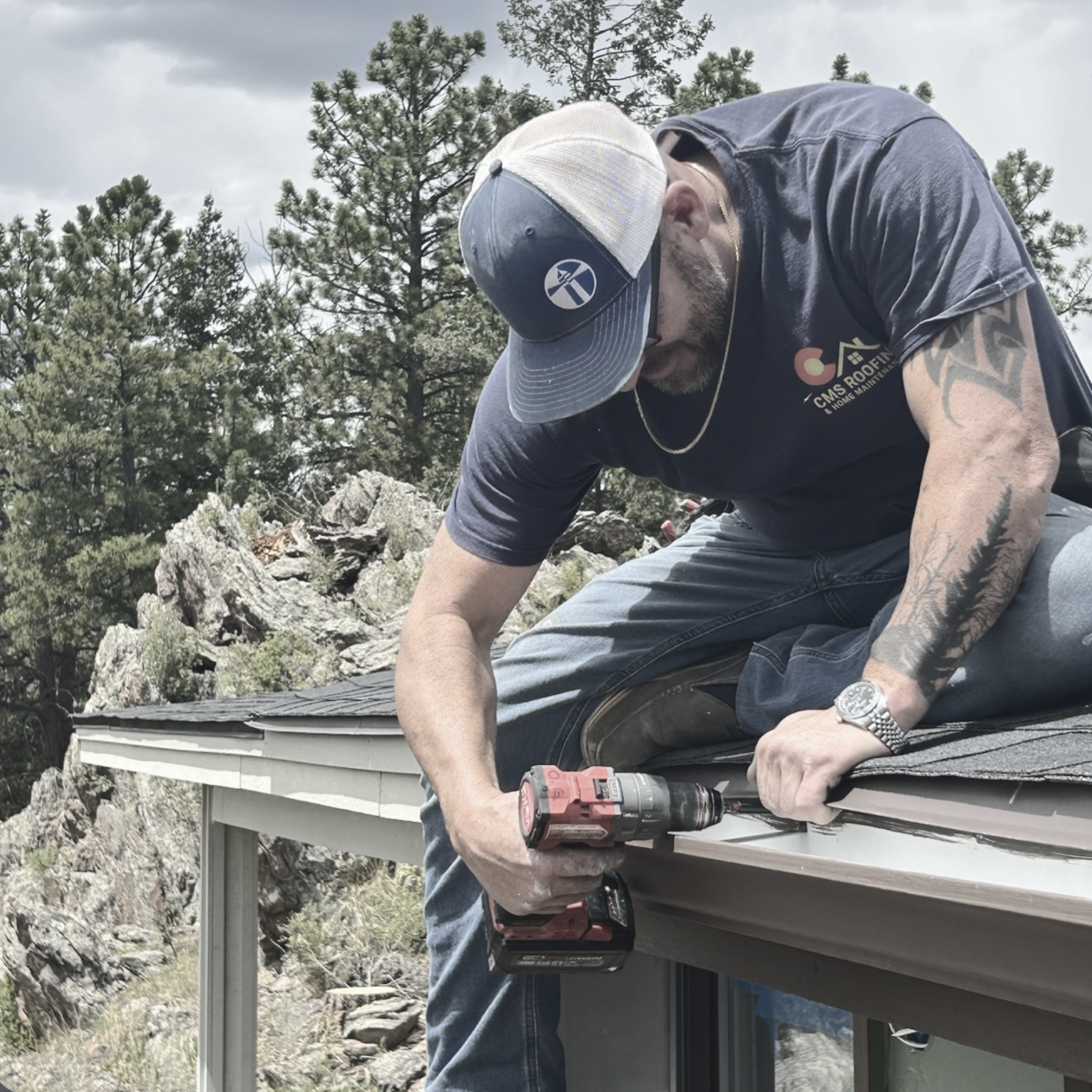 Man using a power drill to install roofing on a house, surrounded by trees and rocky terrain.