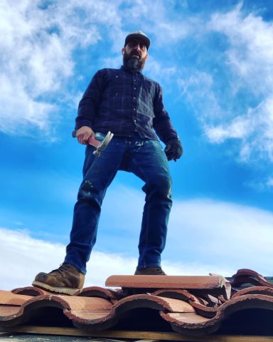 A man stands on a roof with tools, under a blue sky with clouds.